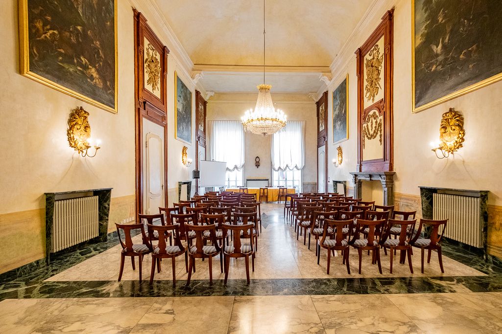 Sala con dipinti alle pareti e lampadario di cristallo, allestita con file di sedie per un incontro