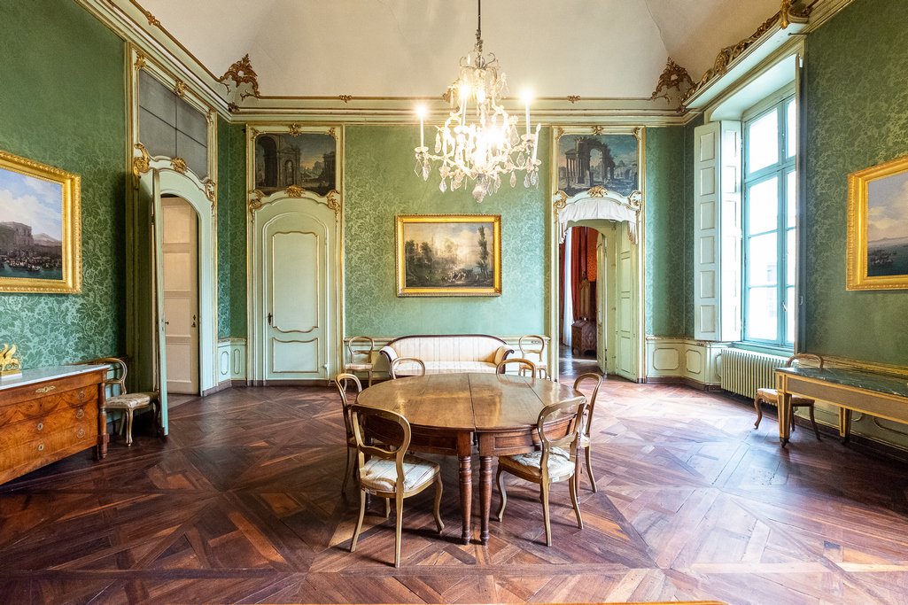 Sala con pareti rivestite di tessuto verde, dipinti in cornici dorate e tavolo rotondo con sedie