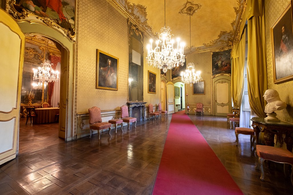 Galleria con passatoia rossa, lampadari di cristallo, ritratti alle pareti e un busto in marmo